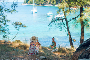 Back view on two young beautiful women caucasian girls friends or sisters taking photos photographing by the trees near the sea water on the seaside summer vacation holiday tourists in sunny day