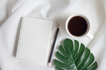 Empty notebook with cup of coffee and monstera leaf on white fabric background. Top view, flat lay.