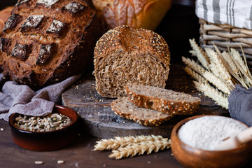 Concept of homemade bread, natural farm products, domestic production. Healthy and tasty organic food. Cut bread, milk, flour. Wheat spica as decor. Closeup, dark wooden background