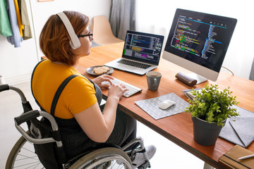 female freelance programmer in modern headphones sitting in wheelchair and using computers while cod
