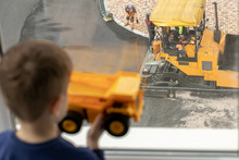 Male Builders Make A Road Near A Residential Complex. The Boy Looks Out The Window And Holds In His Hands A Toy Yellow Car Similar To A Construction One.