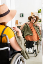 Young Disabled Woman In Hat Sitting In Wheelchair In Front Of Mirror And Trying On Clothes Before Walk