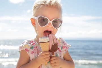 Adorable toddler girl eating ice cream. Portrait of child wearing sunglasses and holding an ice cream with beautiful blue sea and sky behind. Happy summer vacation on the beach.