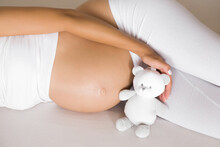 Young Woman Lying Down On Bed In Bedroom. Naked Big Belly Shape. Hand Holding Little Smiling White Teddy Bear For Future Baby. Emotional Loving Moment In Pregnancy Time - 35 Weeks. Baby Expectation.