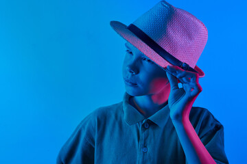 Portrait of a cool boy child stylishly posing in a straw hat on a blue and violet neon light background.