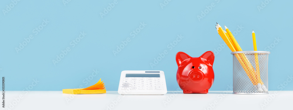 Business or education growth development and success concept banner. White calculator with red piggy coins bank and and yellow pencils on white office or home desk with blue background. Copy space - obrazy, fototapety, plakaty 