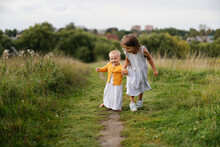 Lovely Little Sisters Walk Together In A Field In The Grass, Children Are Free Countryside, Authentic Lifestyle And Childhood In The Summer.