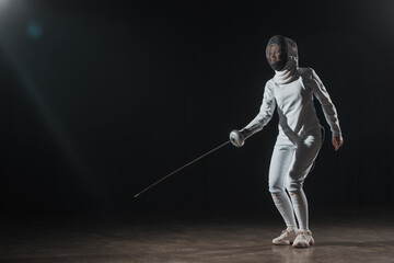 Swordswoman in fencing mask and suit training with rapier under spotlight on black background