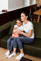 Mother and kid eating popcorn while watching movie on couch at home