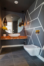 Interior View Of Modern Bathroom With Dark Grey Floor Tiles And Walls With Geometric Pattern, Round Wall Mirror Over Basin On Wooden Railway Sleeper And Wall Mounted Toilet.