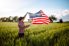 Young Patriotic Farmer Stands Among New Harvest. Boy Walking With The American Flag On The Green Wheat Field Celebrating National Independence Day. 4th Of July Concept.