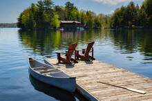 Two Muskoka Chairs Sitting On A Wood Dock Facing A Lake. Across The Calm Water Is A Brown Cottage Nestled Among Green Trees. A Canoe Is Tied To The Dock.