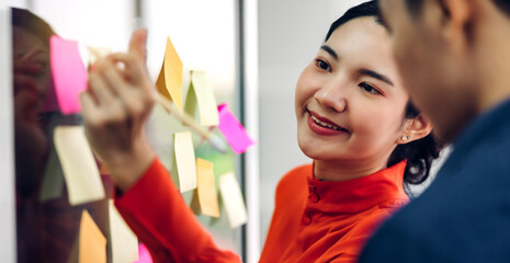 Casual creative happy two smile asian business people planning strategy analysis and brainstorm putting post it stickers note to share idea for startup project on glass board at modern office