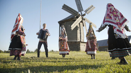 People in traditional russian clothes dancing and singing song outdoor on traditional antique wooden windmill background. Group of happy people wearing national Finno-Ugric clothes.