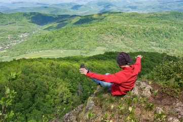 A man in a red jacket sits on the edge of a cliff with his arms outstretched over a beautiful spring landscape. View from above