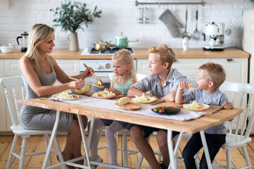 blonde family mother with three kids two sons and daughter eating healthy food in kitchen at home, mom puts green salad on plates to her kids.