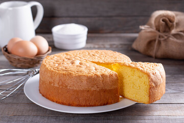 Homemade round sponge cake or chiffon cake on white plate so soft and delicious with ingredients: eggs, flour, milk on wood table