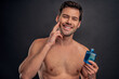 Handsome young bearded man isolated. Portrait of shirtless muscular man is standing on grey background and using face aftershave lotion. Men care concept.