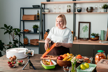 Wall Mural - Happy blond middle aged female looking in smartphone camera while cooking