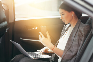 Beautiful woman in a business suit sits in the passenger compartment and checks data through a smartphone and laptop