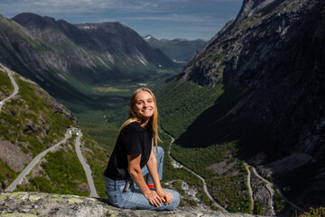 Wall Mural - Trip to Norway. Young blonde woman sits alone over a cliff at Trollstigen road and smiles happy in green valley with high rocky mountains on background in bright summer day