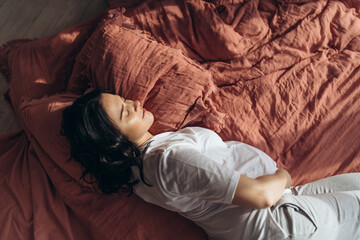 Soft focus. Pregnant young woman sleeping peacefully in bed with one hand on belly. Sun comes through the window. Bed linen of lush lava colour 