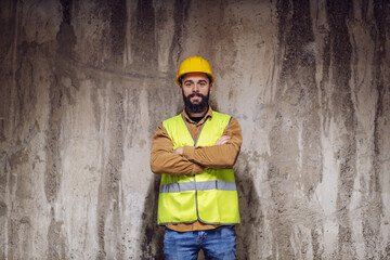 Wall Mural - Young smiling attractive bearded worker in vest with helmet on head standing inside of building in construction process with arms crossed and looking at camera.