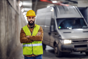 Wall Mural - Young handsome bearded smiling supervisor in vest, with safety helmet on head standing in underground parking lot in construction process with arms crossed. In background is pickup truck.