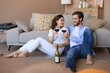 © ty - Happy couple sitting, relaxing on floor in living room, drinking red wine. Smiling young husband and wife rest at home enjoy romantic date on family weekend together