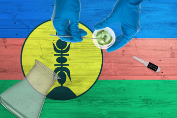 Wall Mural - New Caledonia flag on laboratory table. Medical healthcare technologist holding COVID-19 swab collection kit, wearing blue protective gloves, epidemic concept.