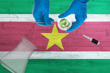 Wall Mural - Suriname flag on laboratory table. Medical healthcare technologist holding COVID-19 swab collection kit, wearing blue protective gloves, epidemic concept.