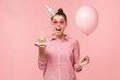© Damir Khabirov - Excited teenage girl celebrating her birthday, wearing hat and glasses, holding balloon, ready to blow candle on cupcake, isolated on pink background
