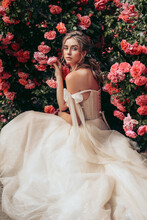 Beautiful Bride With Blond Hair In Luxurious Wedding Dress Posing In The Spring Garden With Blooming Roses Bushes
