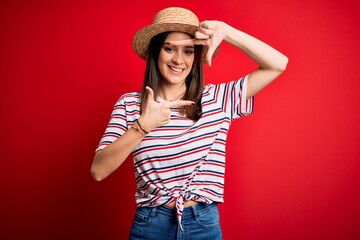 Wall Mural - Young beautiful brunette woman wearing striped t-shirt and summer hat over red background smiling making frame with hands and fingers with happy face. Creativity and photography concept.