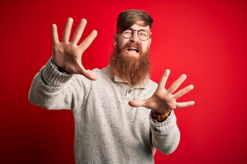 Canvas Print - Handsome Irish redhead man with beard wearing casual sweater and glasses over red background afraid and terrified with fear expression stop gesture with hands, shouting in shock. Panic concept.