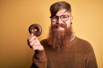 Sticker - Redhead Irish man with beard eating chocolate donut over yellow background with a happy face standing and smiling with a confident smile showing teeth