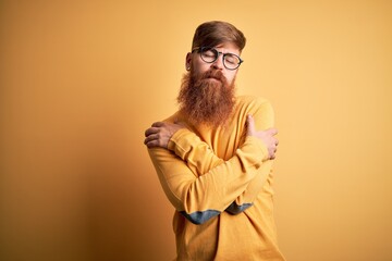 Wall Mural - Handsome Irish redhead man with beard wearing glasses over yellow isolated background Hugging oneself happy and positive, smiling confident. Self love and self care