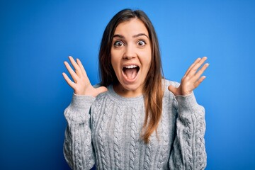 Sticker - Beautiful young woman wearing casual wool sweater standing over blue isolated background celebrating crazy and amazed for success with arms raised and open eyes screaming excited. Winner concept