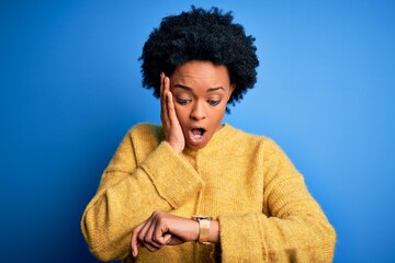 Young beautiful African American afro woman with curly hair wearing yellow casual sweater Looking at the watch time worried, afraid of getting late