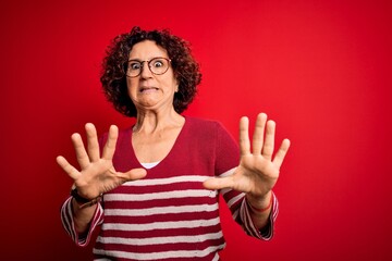 Poster - Middle age beautiful curly hair woman wearing casual striped sweater over red background afraid and terrified with fear expression stop gesture with hands, shouting in shock. Panic concept.