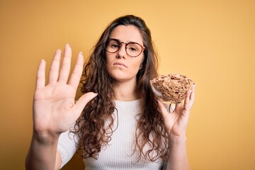 Poster - Young beautiful woman with curly hair holding bowl with healthy corn flakes cereals with open hand doing stop sign with serious and confident expression, defense gesture