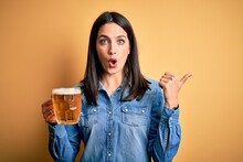 Young Woman With Blue Eyes Drinking Jar Of Beer Standing Over Isolated Yellow Background Surprised Pointing With Hand Finger To The Side, Open Mouth Amazed Expression.