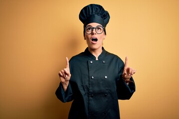 Canvas Print - Young beautiful brunette chef woman wearing cooker uniform and hat over yellow background amazed and surprised looking up and pointing with fingers and raised arms.