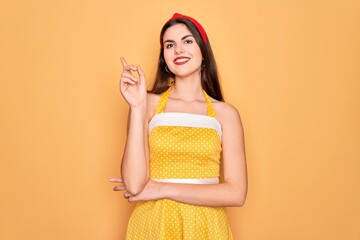 Poster - Young beautiful pin up woman wearing 50s fashion vintage dress over yellow background with a big smile on face, pointing with hand and finger to the side looking at the camera.