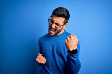 young handsome man with beard wearing casual sweater and glasses over blue background celebrating su