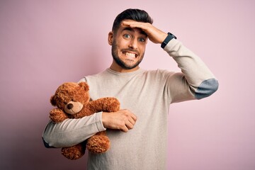 Sticker - Young handsome man holding teddy bear standing over isolated pink background stressed with hand on head, shocked with shame and surprise face, angry and frustrated. Fear and upset for mistake.