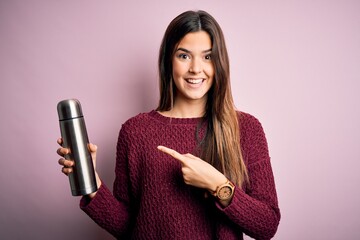 Sticker - Young beautiful girl holding thermo with water standing over isolated pink background very happy pointing with hand and finger