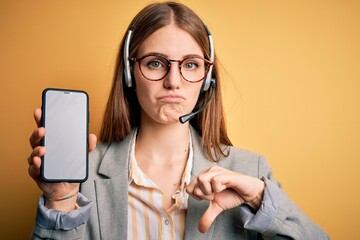 Wall Mural - Young redhead call center agent woman using headset holding showing screen of smartphone with angry face, negative sign showing dislike with thumbs down, rejection concept