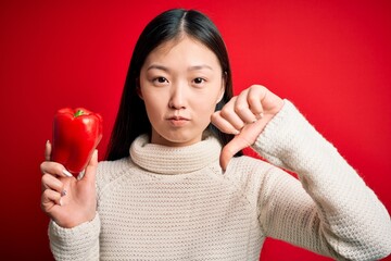 Poster - Young asian woman holding healthy and fresh red pepper over isolated background with angry face, negative sign showing dislike with thumbs down, rejection concept