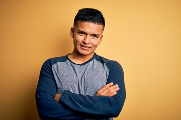 Young handsome latin man wearing casual t-shirt standing over yellow background happy face smiling with crossed arms looking at the camera. Positive person.